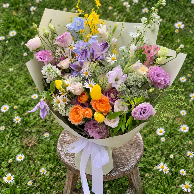 Spring Blooms bouquet with purple, yellow and orange flowers, pale green wrapping and a white ribbon on a wooden stool in a daisy meadow